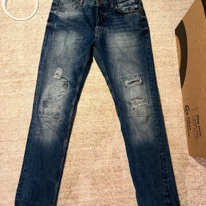 Blå jeans från Adrian Hammond - Säljer ett par snygga och unika blå jeans från Adrian Hammond Hollywood i storlek W32 men sitter mer som en 31. passformen är straight fast mer åt slim hållet. De har en cool slitning på framsidan och en klassisk femficksdesign. Perfekta för en avslappnad stil eller en utekväll. Tveka inte på att höra av er för frågor och funderingar:)