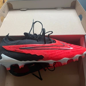 Nike fotbollsskor i röd och svart - Säljer ett par grymma Nike fotbollsskor i röd och svart. De har en snygg design med vita detaljer och är perfekta för fotbollsplanen. Skorna har snörning och en robust sula med dobbar för bästa grepp. Perfekt för dig som vill dominera på planen! De är använda max 1-2 gånger och är i väldigt bra skick, box ingår men ej påse! Hojta till vid frågor eller funderingar🙌