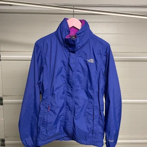 Blå vindjacka från The North Face - Säljer en blå vindjacka från The North Face i mycket bra skick. Jackan har en dragkedja framtill och är långärmad. Perfekt för vår och höst med sin lätta och skyddande design. Den har en snygg rosa insida som ger en fin kontrast.Säljes i befintligt skick. Storlek: L