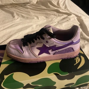 Bapesta dunks - Snygga lila sneakers från Bape med en stjärndetalj på sidan. Skorna har svarta snören och kommer i en cool kamouflagemönstrad låda. Perfekta för att ge din stil en unik touch.