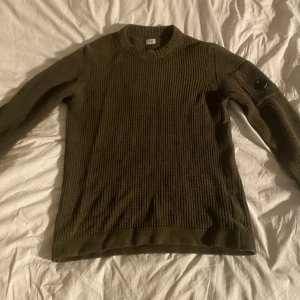 Olivgrön stickad tröja från C.P. Company - Säljer en olivgrön stickad tröja från C.P. Company. KNAPPT ANVÄND!Tröjan har en rund halsringning och långa ärmar. Den är dekorerad med en svart detalj på ärmen med C.P. Companys logotyp. Perfekt för kyliga dagar.