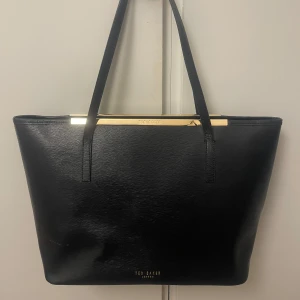 Ted Baker väska - Svart axelväska från Ted Baker med gulddetaljer upptill. Inga slitage på handtagen. På bilden reflekterar ljuset lite på väskan, det är ingen avfärgning.