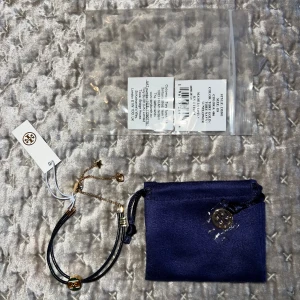 Svart och guld armband med logga - Snyggt armband med svart band och gulddetaljer. Armbandet har en rund logga i mitten med gröna och guldiga ränder. Perfekt accessoar för att ge din outfit en lyxig touch. Kommer med en blå förvaringspåse.