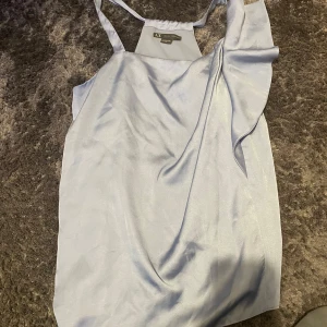 Ljusblå klänning från Armani Exchange - Säljer en elegant ljusblå blus från Armani Exchange i sidenliknande material. Blusen har smala axelband och en fin volangdetalj på framsidan. Perfekt för sommarens fester eller en speciell kväll. 🩵