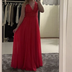 Röd/rosa balklänning med spetsdetaljer - Säljer en  röd/rosa långklänning med vackra spetsdetaljer och djup v-ringning. Klänningen har en elegant öppen rygg och är perfekt för festliga tillfällen. Materialet är mjukt och flödande, vilket ger en fin passform. Perfekt för bal!🥳