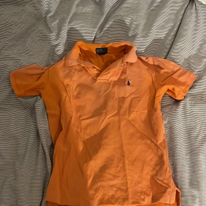 Orange kortärmad pikétröja från Polo Ralph Lauren - Säljer en snygg orange pikétröja från Polo Ralph Lauren i storlek M. Tröjan har korta ärmar och en klassisk krage med knappar. Den är broderad med det ikoniska Polo-loggan i blått på bröstet. Perfekt för en avslappnad men stilren look!