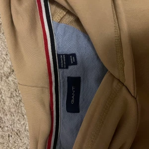 Beige hoodie från GANT - Säljer en snygg beige hoodie från GANT i storlek M. Den är i bra skick och perfekt för höst och vår. Hoodien har en klassisk känguruficka och en liten broderad logga på bröstet. Superbekväm och stilren för vardagsbruk!