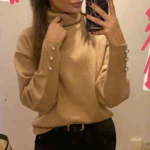 Beige polotröja med pärldetaljer - Säljer en stilren beige polotröja med långa ärmar och dekorativa pärlknappar vid ärmsluten. Perfekt för kyligare dagar och ger en elegant touch till din outfit.