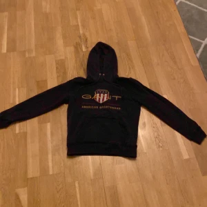 Svart hoodie från GANT - Säljer en svart hoodie från GANT med broderad logga och texten 'American Sportswear' på framsidan. Hoodien har en klassisk design med justerbar huva och långa ärmar. Perfekt för en avslappnad stil.