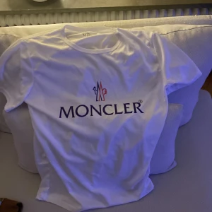 Vit t-shirt MONCLER - Säljer en snygg vit t-shirt från Moncler i bomull. Den har korta ärmar och en klassisk rund halsringning. Moncler-loggan är tryckt i svart på framsidan. Perfekt för en stilren och avslappnad look!