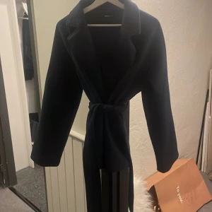 Mörkblå kappa med knytband - Säljer en elegant mörkblå kappa i mycket bra skick. Den har ett knytband i midjan och långa ärmar, perfekt för höst och vår. Stilren och tidlös design som passar till många olika tillfällen.