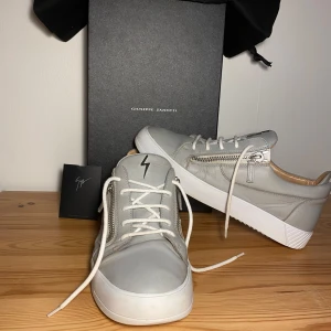Grå sneakers från Giuseppe Zanotti - Stilrena grå sneakers från Giuseppe Zanotti med snörning och dragkedja på sidan. Skorna har en vit sula och en diskret blixtdetalj på plösen. Perfekta för en modern och avslappnad look. Allt OG medföljer