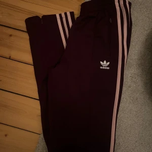 Lila  träningsbyxor från Adidas - Snygga mörklila träningsbyxor från Adidas med de klassiska tre ränderna i rosa längs sidorna. Byxorna har en elastisk midja och dragkedja vid ankel för extra funktionalitet. Perfekta för en sportig look!