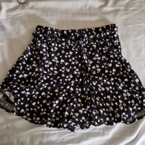 Svart kjol med blommigt mönster - Säljer en gullig kjol, svarta med vitt blommigt mönster. Har ett par inbyggda short. Kjolen har en elastisk midja med knytband för extra justering. De är gjorda i ett lätt och luftigt material, vilket gör dem idealiska för både vardag och fest.