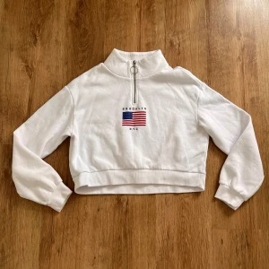 Snygg Vit croppad tröja från H&M Divided - Säljer en superfin vit croppad tröja från H&M Divided. Den har en halv dragkedja framtill och ett tryck med texten 'Brooklyn NYC' och en amerikansk flagga. Tröjan är långärmad och gjord av en mjuk bomullsblandning, perfekt för en casual look. Passar perfekt till höst och vår!