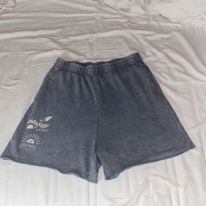 Grå shorts med tryck - Säljer ett par grå shorts med elastisk midja och coolt tryck på ena benet. Perfekta för en avslappnad dag eller en tur till stranden. De är i bra skick och superbekväma!