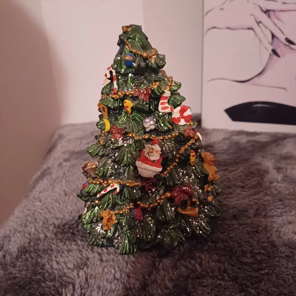 Säljer en superfin julgranformad dekoration med massor av detaljer. Den är grön med små färgglada dekorationer som tomtar, godis och glitter. Perfekt för att skapa julstämning hemma! Den är gjord av ett hårt material och har en öppning i botten, kanske för att sätta in ett ljus eller liknande. Perfekt för julpyntet! Den snurrar själv och låter🎄✨. Accessoarer.