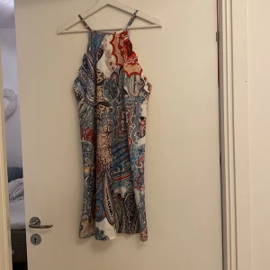 Färgglad mönstrad sommarklänning - Säljer en superfin och färgglad sommarklänning med ett unikt paisleymönster i olika färger som blå, röd och beige. Klänningen är ärmlös och har smala axelband, perfekt för varma sommardagar. Materialet känns lätt och luftigt, vilket gör den idealisk för både strand och fest. Passar perfekt för dig som vill sticka ut med en bohemisk stil!