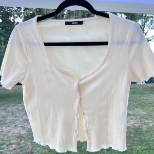 Gul ribbad topp med knappar - En ljusgul ribbad topp med knappar från Bikbok. Inköpt för flera år sedan men använd max 3 gånger 💕💕 Fin att använda för sig själv eller öppen med en liten topp under 🥰  Längd: 45 cm Axel: 35 cm Byst: 75 cm