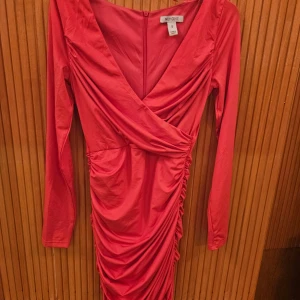 Röd omlottklänning från New One - Säljer en superfin rosa omlottklänning från Nelly i storlek S. Klänningen har en v-ringad design och långa ärmar, perfekt för festliga tillfällen. Materialet känns mjukt och bekvämt, och den har en snygg drapering på sidan som ger en extra touch. 