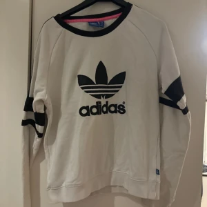 Vit och svart tröja från Adidas - Säljer en snygg vit tröja från Adidas med deras klassiska logga i svart på framsidan. Tröjan har långa ärmar med svarta detaljer vid ärmsluten och en svart krage. Perfekt för en sportig och avslappnad stil!