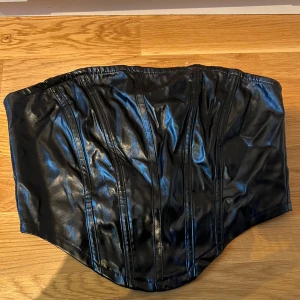 Svart skinnkorsett - Säljer en snygg svart skinn topp med dragkedja i ryggen. Toppen har en glansig finish och ger en tight passform.