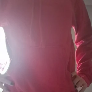 Rosa hoodie - Säljer en supermysig röd hoodie som är perfekt för chilliga dagar. Den har en klassisk passform med en stor ficka fram och en skön huva med dragsko. Materialet känns mjukt och bekvämt, perfekt för både hemma och ute.