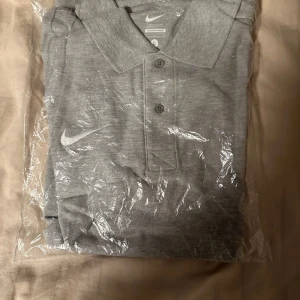 Grå pikétröja från Nike - Säljer en snygg och stilren grå pikétröja från Nike. Perfekt för både vardag och träning. Tröjan har en klassisk krage och knappar framtill. Den är helt ny och ligger fortfarande i förpackningen. Passar perfekt för en sportig look! Storlek L passar M