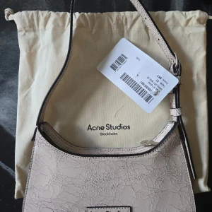 Axelväska från Acne Studios - Aldrig använd Acne Studios Platt Micro axelväska i pastel pink. Väskan har en unik krackelerad yta som ger den en cool och annorlunda look. Den är gjord i skinn och har en praktisk dragkedja upptill. Nypris 8300kr.