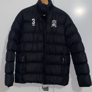 Svart dunjacka med broderad logga - Säljer en svart dunjacka med en cool broderad logga på bröstet och siffran 3. Jackan har en dragkedja framtill och är perfekt för kalla dagar. Den är i bra skick och supervarm!