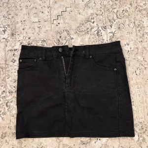Svart jeanskjol - Säljer en snygg svart jeanskjol i kort modell. Kjolen har en klassisk design med dragkedja och knapp framtill samt fickor på sidorna. Perfekt för både vardag och fest! 🖤