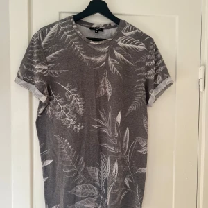 Grå t-shirt med bladmönster från New Look - Säljer en snygg grå t-shirt från New Look med ett coolt bladmönster i vitt. Perfekt för en avslappnad stil, den har korta ärmar och är gjord i ett mjukt material. Passar perfekt till både jeans och shorts!