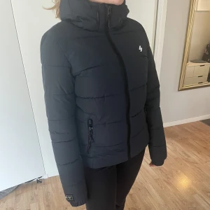 Superdry jacka puffer bra skick - Vadderad pufferjacka Mörkblå • Mjukt fleecefoder •huva med dragsko • Två dragkedjefickor • En innerficka i mesh • Resår i ärmsluten • Dragsko i nederkanten  Märkt XS, men rymlig i storleken. Passar även S/M. Jättefint skick, endast använd enstaka gånger.