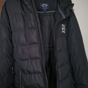 Svart pufferjacka från Vinson - Säljer en svart pufferjacka från Vinson i mycket bra skick. Jackan har en dragkedja framtill och en broderad logga på bröstet. Perfekt för kalla dagar med sin quiltade design och långa ärmar. Den är både stilren och praktisk för vintern.