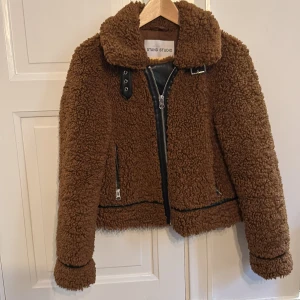 Teddyjacka från Stand Studio - Säljer en supermysig brun teddyjacka i aviotor stil, från Stand Studio. Den har en cool dragkedja framtill och snygga detaljer med bälte vid kragen. Perfekt för höst och vinter när du vill hålla dig varm och stilren. Jackan är i mycket bra skick och redo för nya äventyr! Fickor på båd sidor. Silvriga dragkedjor/spännen.