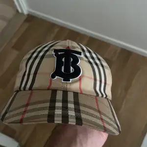 Säljer en snygg rutig keps från Burberry i beige, svart och röd. Den har ett stort broderat 'B' framtill och är perfekt för både vardag och fest. Kepsen är i mycket bra skick och har en klassisk design som aldrig går ur stil. Kepsen är äkta o köpt från NK.