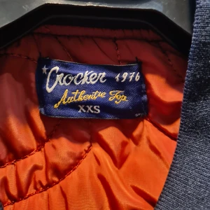 Mörkblå bomberjacka från Crocker - Säljer en snygg mörkblå bomberjacka från Crocker i storlek XXS. Jackan har en cool orange insida och dragkedja framtill. Perfekt för höst och vår, med en klassisk bomberstil och ribbade muddar vid ärmslut och nederkant. Passar både till vardags och fest! 