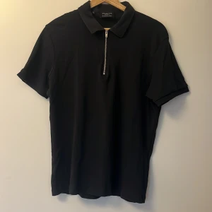 Svart pikétröja från Selected Homme - En snygg svart pikétröja från Selected Homme i storlek M, säljes då den inte riktigt är min stil längre. Tröjan har en dragkedja framtill och korta ärmar, perfekt för en avslappnad men ändå uppklädd look. 