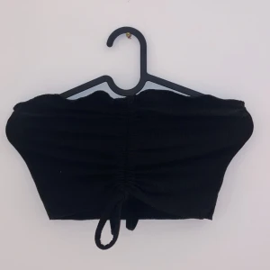 Svart tubtopp - Snygg svart tubtopp i ett stretchigt material. Perfekt för sommarens fester eller en dag på stranden. Toppen har en knytning framtill som ger en fin detalj och gör den justerbar. Passar bra till både kjol och jeans för en avslappnad men stilren look.