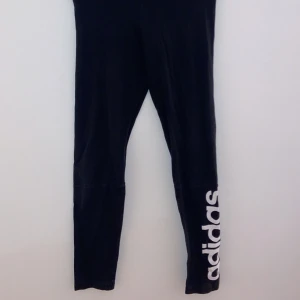 Svarta leggings från Adidas - Säljer ett par svarta leggings från Adidas i bra skick. De har en vit logga längs benet och är perfekta för träning eller en avslappnad dag. Materialet är mjukt och stretchigt för bästa komfort. Passar perfekt till både gymmet och vardagsoutfiten!