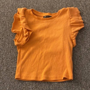 Orange ribbad topp med volangärmar från Zara - Säljer en superfin orange ribbad topp från Zara. Den har korta ärmar med volangdetaljer som ger en söt touch. Perfekt för sommardagar eller en avslappnad fest. Toppen är i bra skick och redo för en ny ägare!