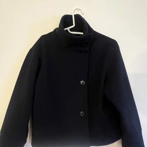 Svart kappa från Gina Tricot - Säljer en stilren svart kappa från Gina Tricot i storlek XXS. Den har en hög krage och knäppning framtill med stora knappar. Perfekt för höst och vinter, och ger en elegant look till vilken outfit som helst. 🖤