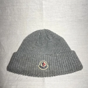 Grå mössa från Moncler - Säljer en snygg grå ribbad mössa från Moncler. Den är perfekt för kalla dagar och har en klassisk design med Moncler-loggan framtill. Mössan är i mycket bra skick och passar perfekt till både vardag och vinteraktiviteter.