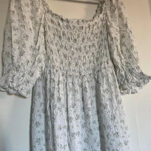 Blommig långklänning med puffärmar - Superfin långklänning i vitt med lila blommönster. Den har puffiga trekvartsärmar och en smockad överdel som ger en romantisk look. Perfekt för vår och sommar, kanske till en midsommarfest! Materialet känns lätt och luftigt.