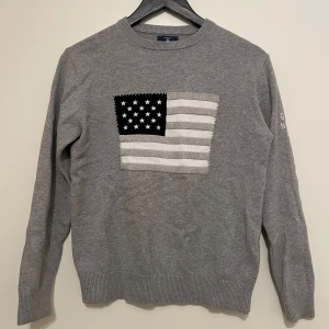 Grå tröja från GANT med flaggmotiv - Säljer en snygg grå tröja från GANT med ett coolt flaggmotiv på framsidan. Tröjan har långa ärmar och är i ett mjukt material som känns skönt mot huden. Perfekt för både höst och vinter. Detaljerad med broderad text på ärmen. Passar perfekt för en casual look!