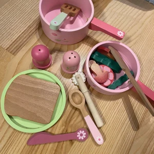 Rosa leksaksköksset i trä - Ett gulligt rosa leksaksköksset i trä med kastruller, stekspade, sked och tallrik. Setet inkluderar även salt- och pepparkar samt olika leksaksmatbitar. Perfekt för små kockar som älskar att laga mat i sitt eget lilla kök. Färgglada detaljer och blommönster gör det extra charmigt.