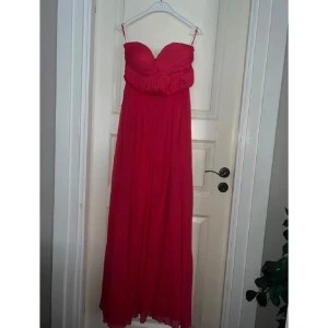 Röd axelbandslös långklänning - Säljer en fantastisk röd axelbandslös långklänning från Gina Tricot. Klänningen har en vacker drapering över bysten och är gjord i ett mjukt, flödande material som ger en elegant look. Perfekt för festliga tillfällen eller en somrig bal. Den är i mycket bra skick och redo att bäras av någon som vill känna sig extra speciell.