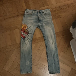 Jeans med tryck från Palm Angels - Säljer ett par coola jeans från Palm Angels med ett unikt tryck av en brinnande mun på ena benet. De är ljusblå och är i bra skick 