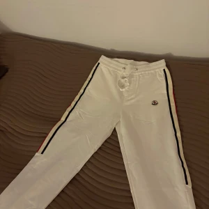 Vita mjukisbyxor från Moncler - Säljer ett par vita mjukisbyxor från Moncler i nytt skick. De har en snygg snörning i midjan och ränder i blått och rött längs sidorna. Perfekta för en avslappnad stil eller en dag hemma. Moncler-loggan är broderad på framsidan. Superbekväma och stilrena!