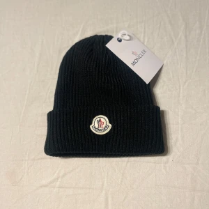Svart mössa från Moncler - Säljer en snygg svart ribbad mössa från Moncler i oanvänt skick. Den är perfekt för vintern och har en broderad Moncler-logga framtill. Mössan är gjord i ett mjukt och varmt material, vilket gör den idealisk för kalla dagar. Passar både till vardags och mer uppklädda tillfällen.